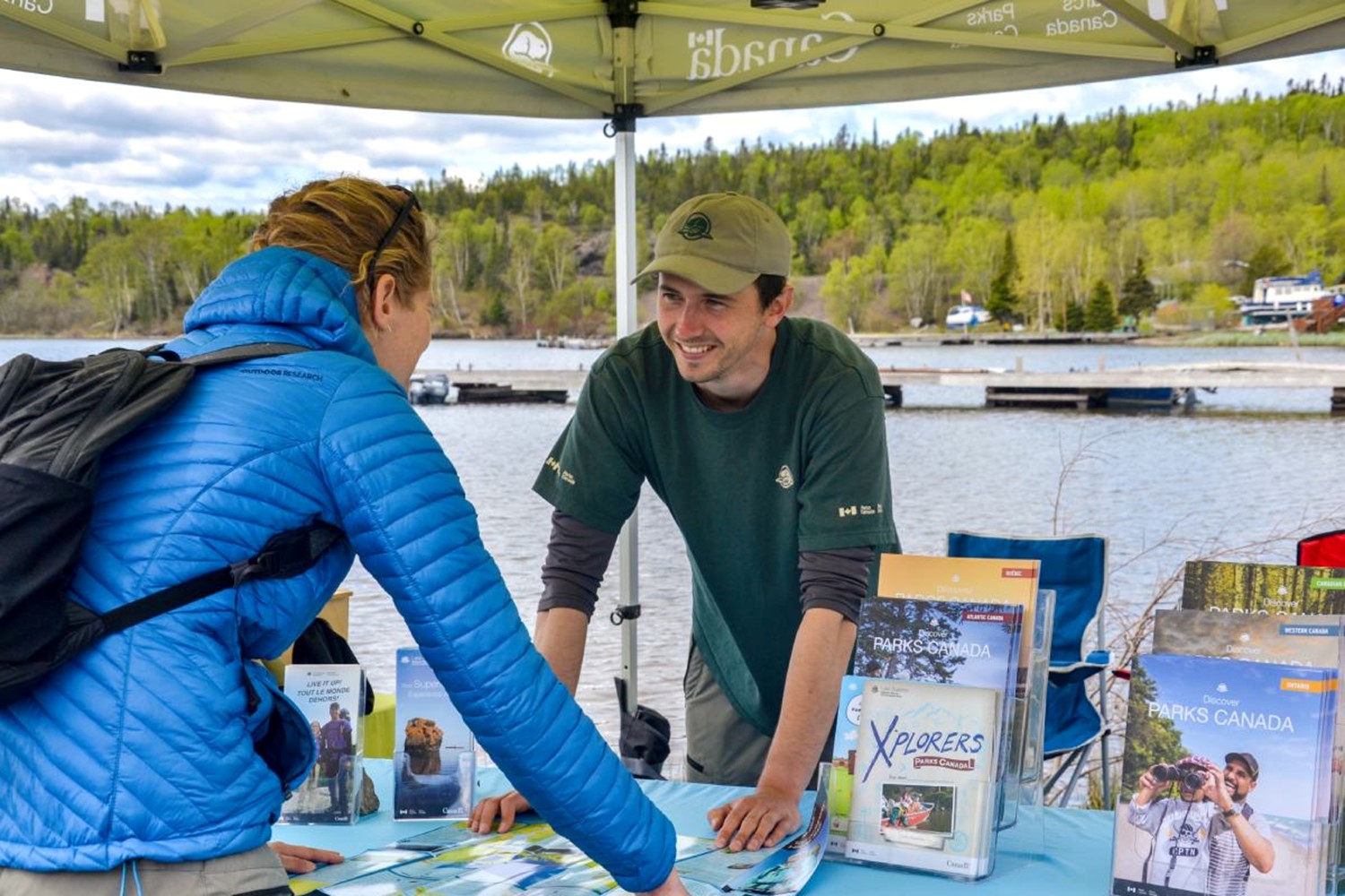 Parks Canada representative sharing information with a visitor at a booth, with water and trees in the background.