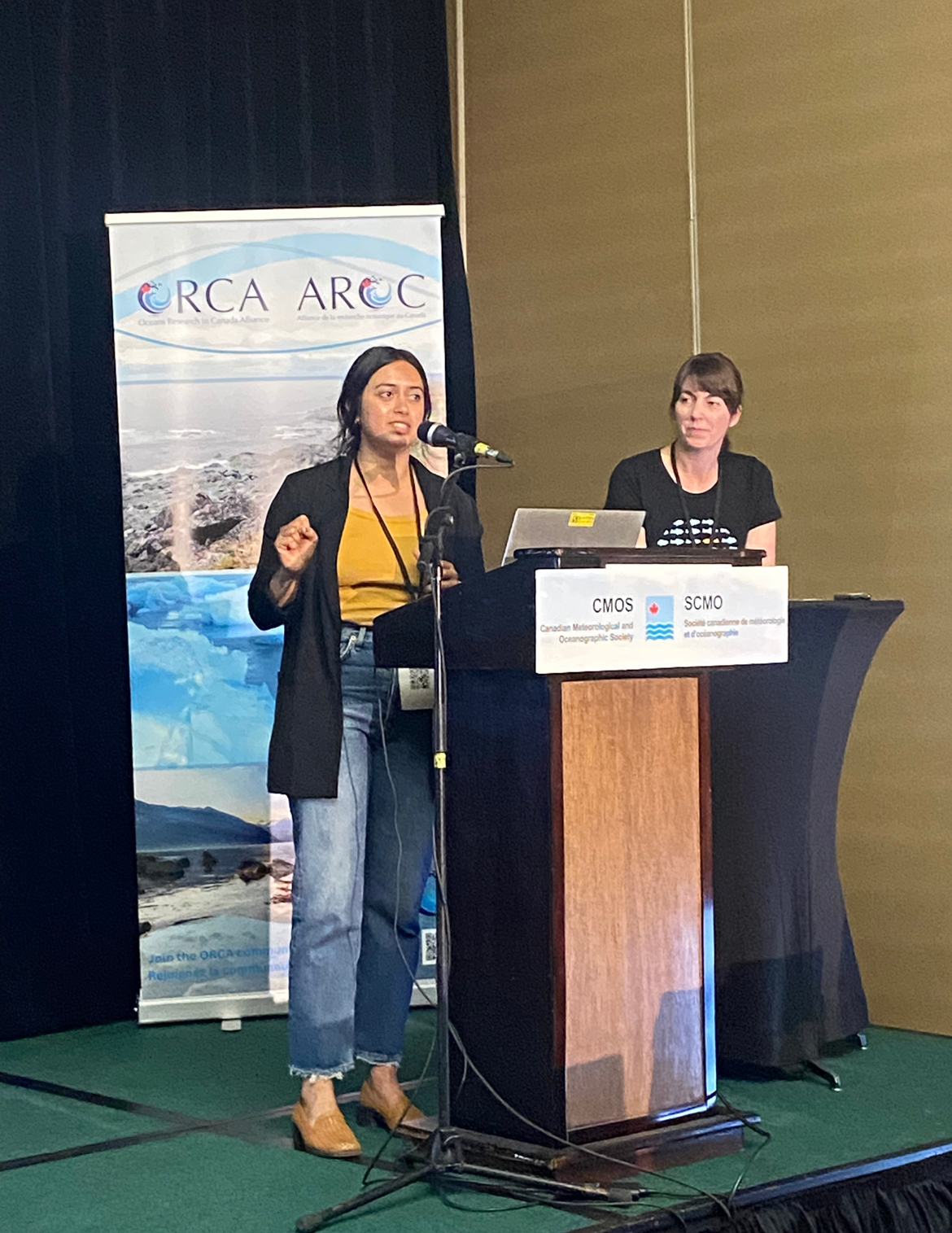 Two women stand at the podium. Neha Acharya-Patel is mid-speech at the microphone, while Andi White is standing to her left. Behind them, there is an ORCA-AROC banner with ocean images.