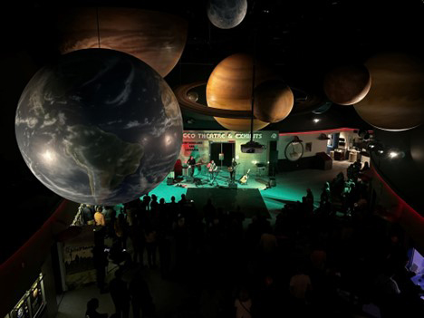 Groupe sur scène jouant pour un grand public au Johnson Geo Centre, où des répliques de planètes sont suspendues au plafond d’une salle peu éclairée.