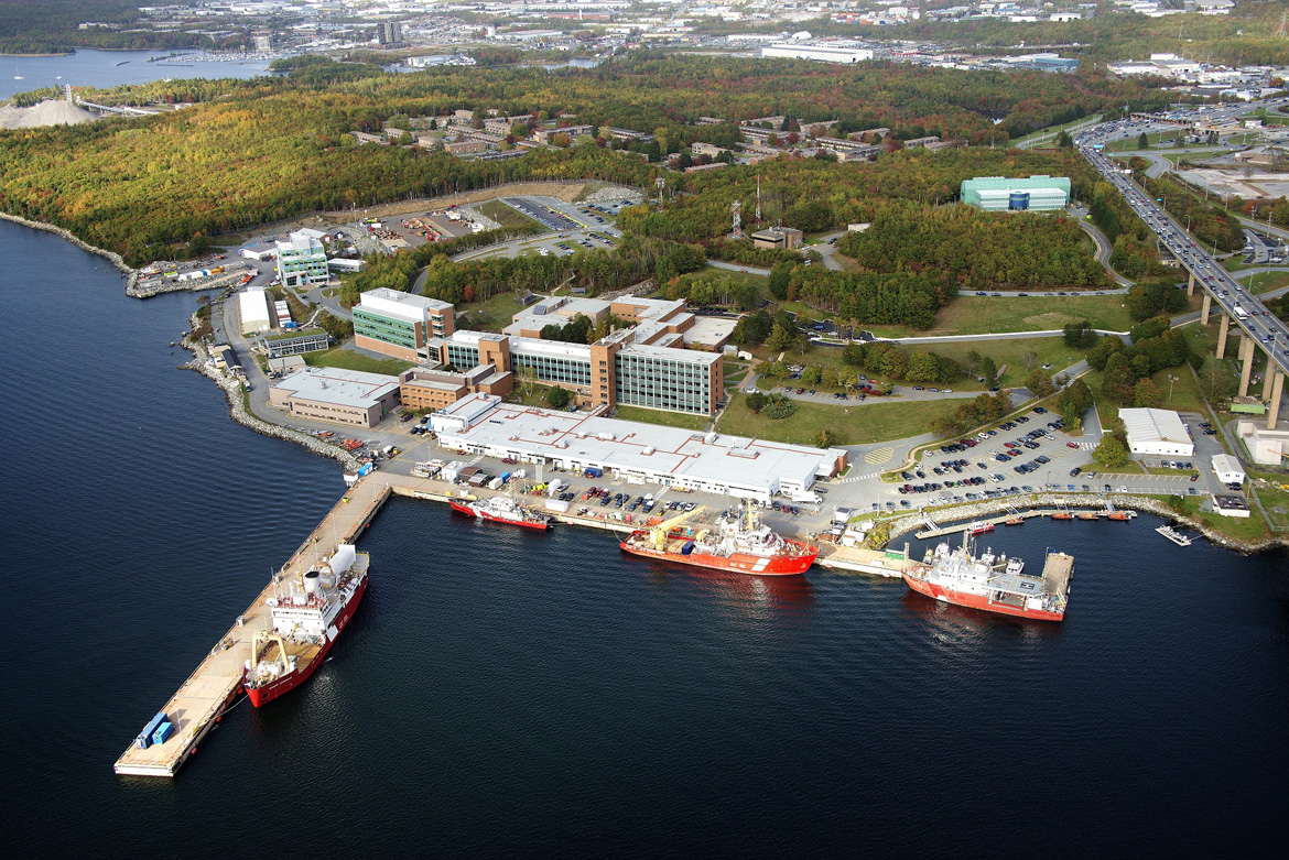 Vue aérienne d'un certain nombre d'immeubles de bureaux à côté d'une jetée avec des navires de la Garde côtière canadienne. Les immeubles de bureaux sont entourés d'une forêt.